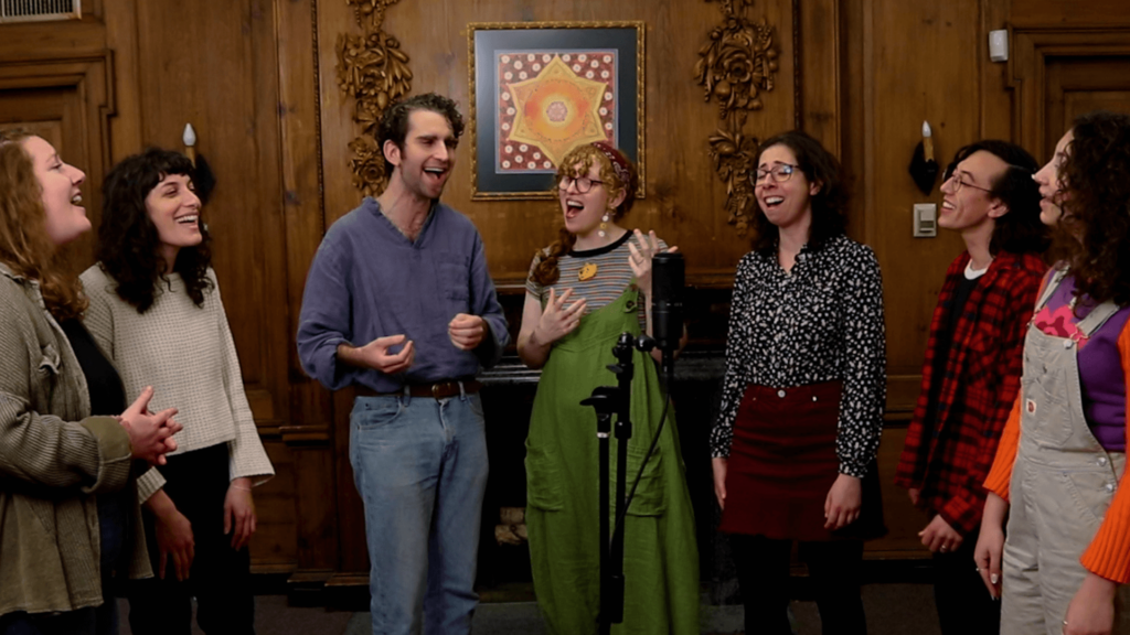 Seven members of the RRC Music Collective singing together in a room with wood paneling and ornate wall decorations.