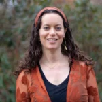 Woman with curly hair and a headband, wearing an orange floral top, smiles at the camera in a wooded area.