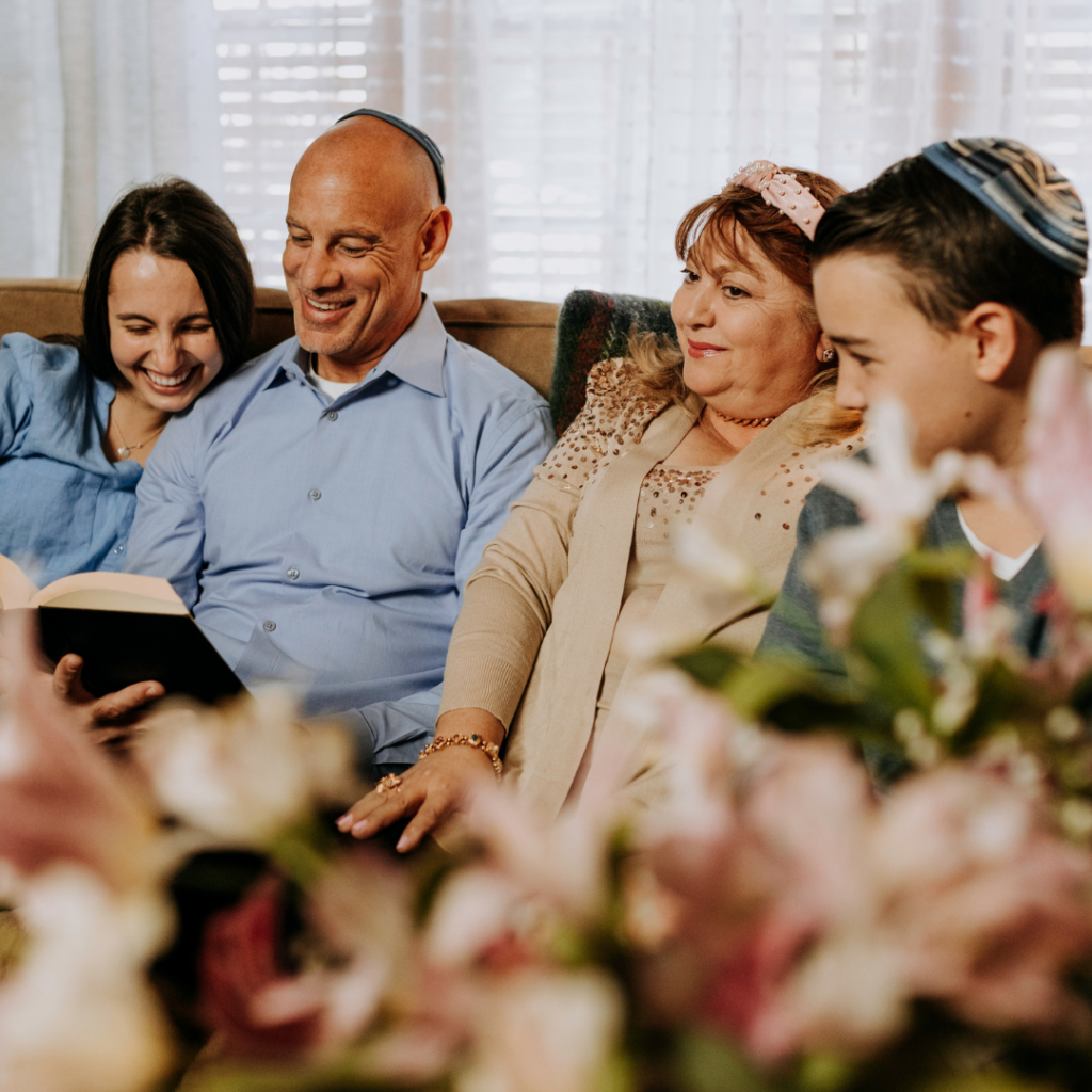 Family of four sitting on a couch, smiling and reading together, with flowers in the foreground.