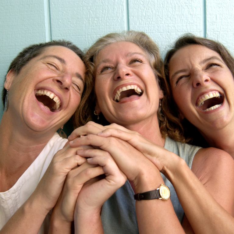 Three women laughing joyfully, holding hands, against a light blue wall background.