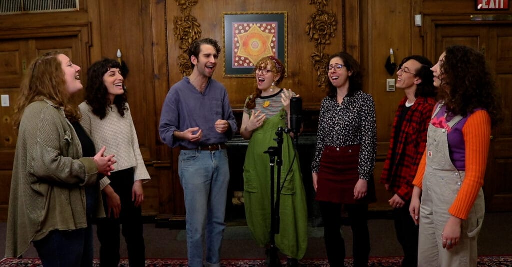 Seven people standing in a semi-circle, joyfully singing in a wood-paneled room with a decorative wall hanging.