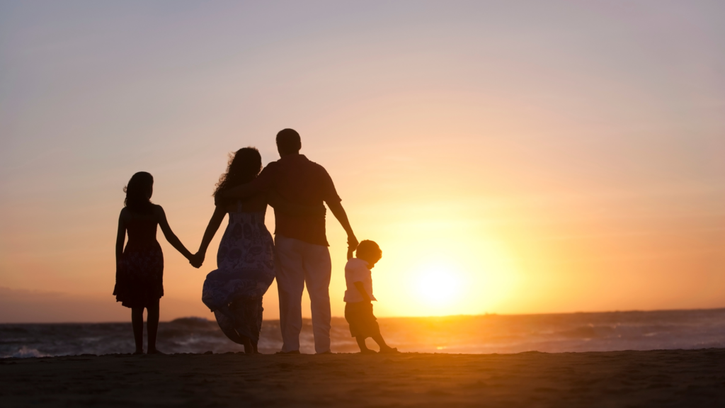 Silhouetted family holding hands, watching a beach sunset together.