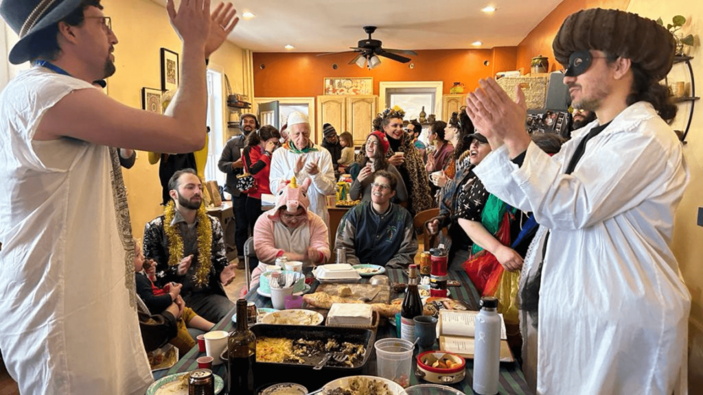 A lively Purim party with people in vibrant outfits, clapping and cheering around a dining table.