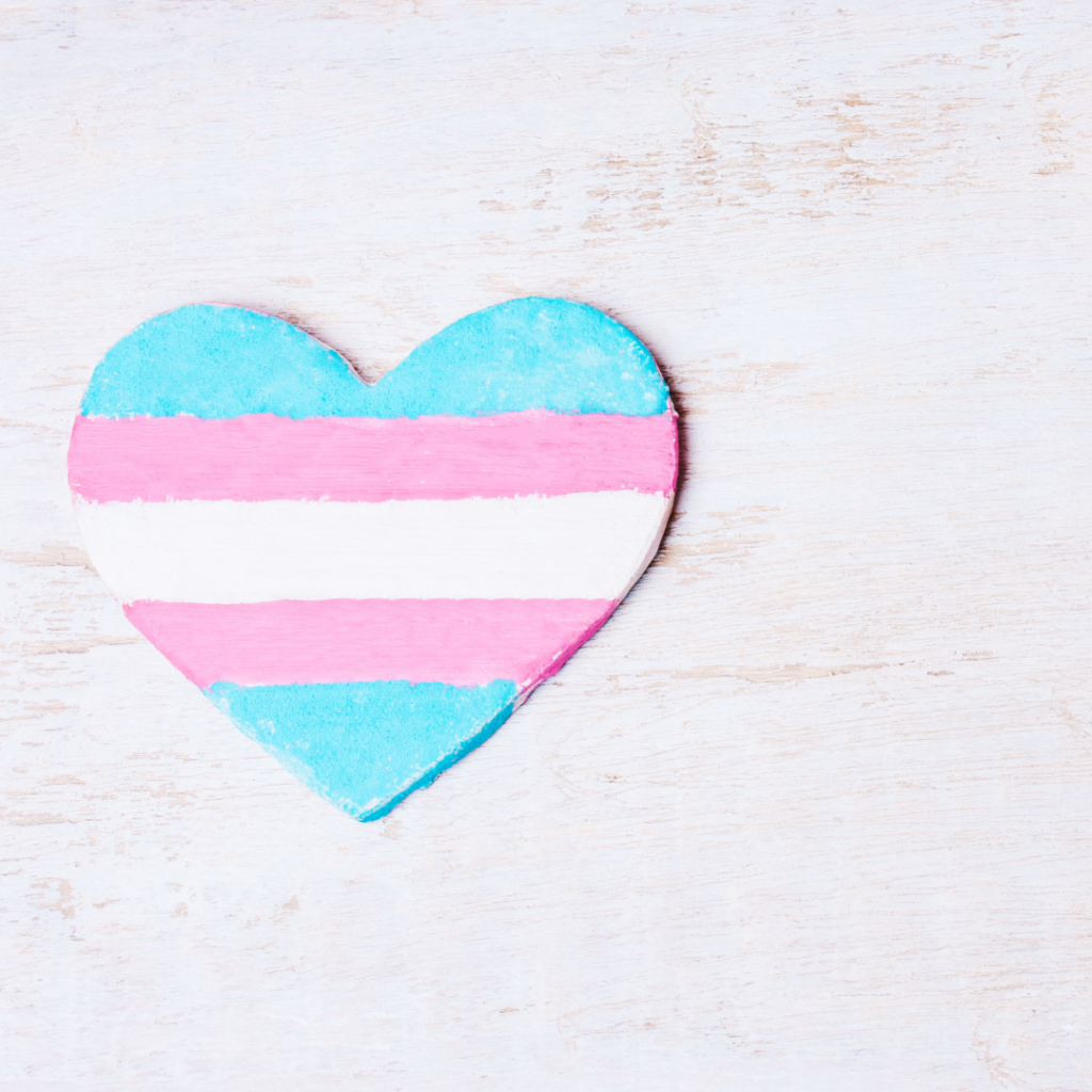 Heart-shaped object painted with transgender pride colors on a light wooden surface.