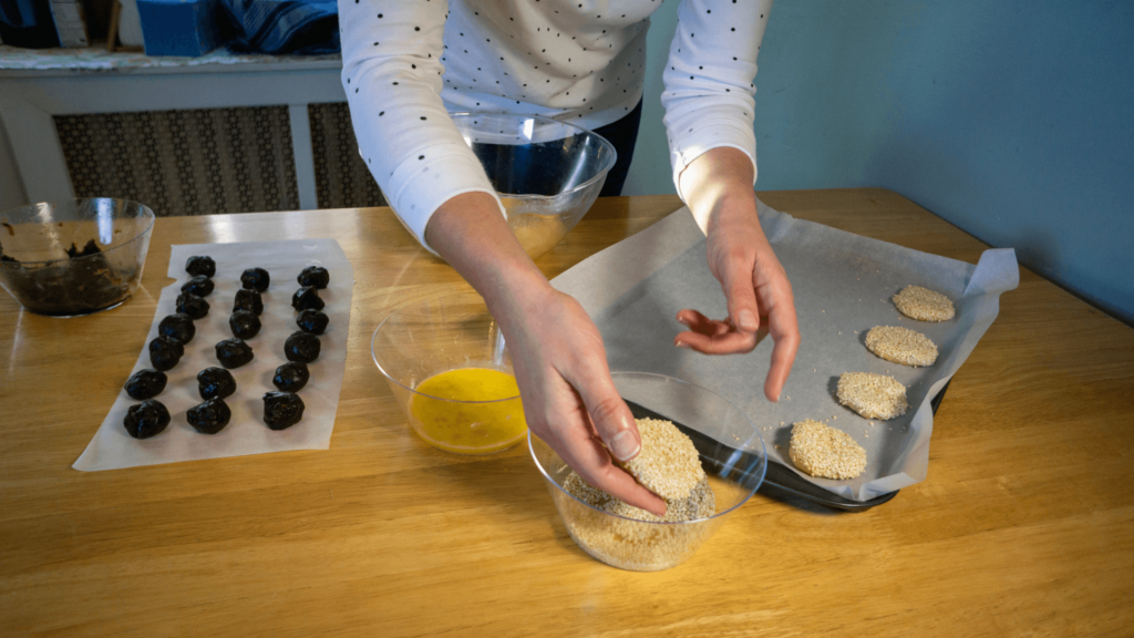 Person preparing dough with sesame seeds on a tray, with a bowl of egg wash and lined balls on the table.