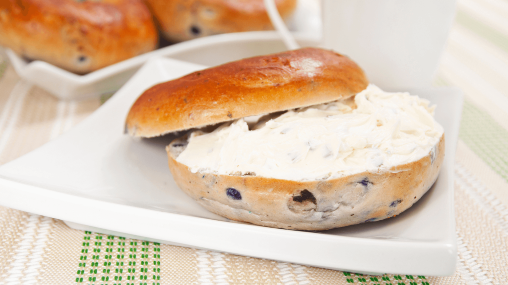 Blueberry bagel with cream cheese on a white plate, placed on a striped tablecloth.