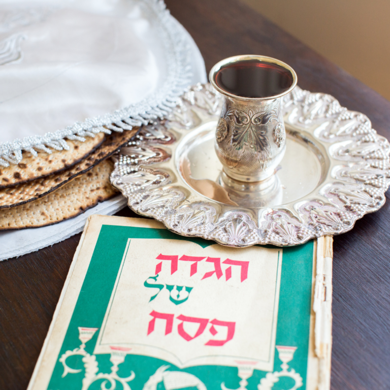 Silver cup on a decorative plate, matzah, and a Haggadah book on a wooden table.