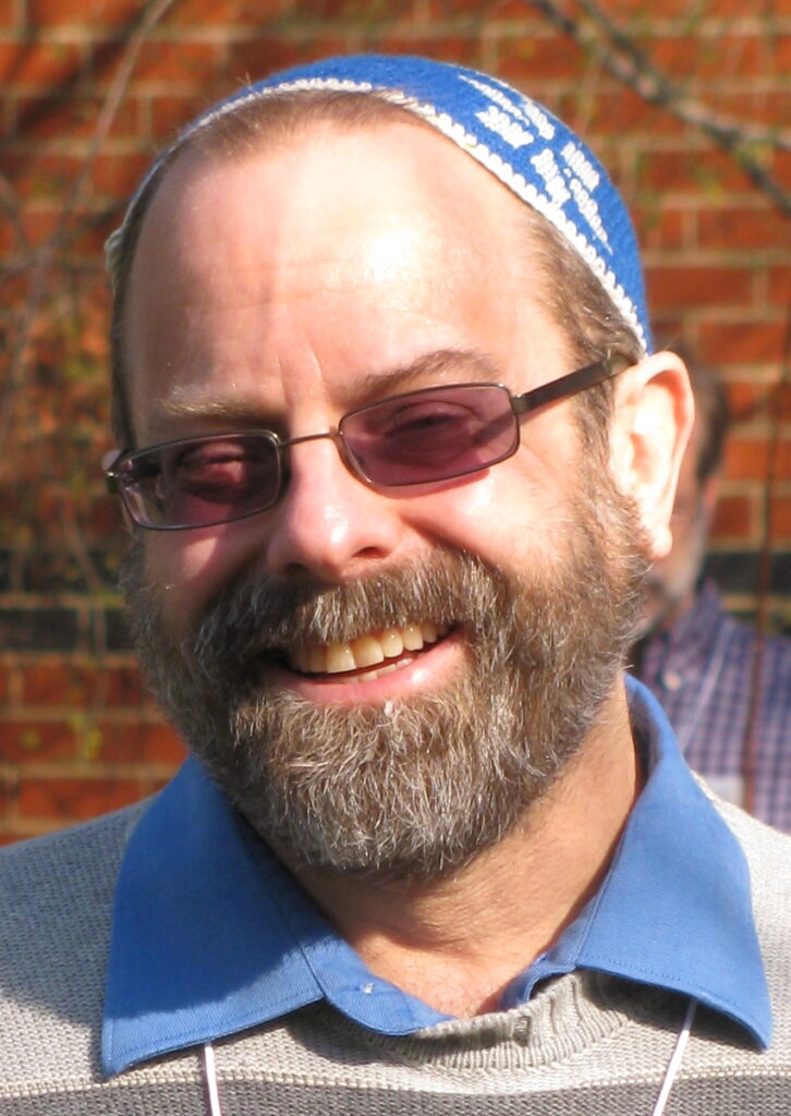 A smiling man with a beard, glasses, and a blue patterned cap, wearing a blue collared shirt outdoors.