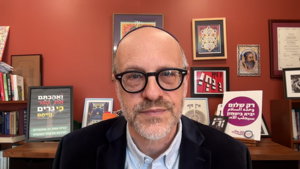 Man with glasses and a kippah in an office, posters with Hebrew text on the wall behind him.