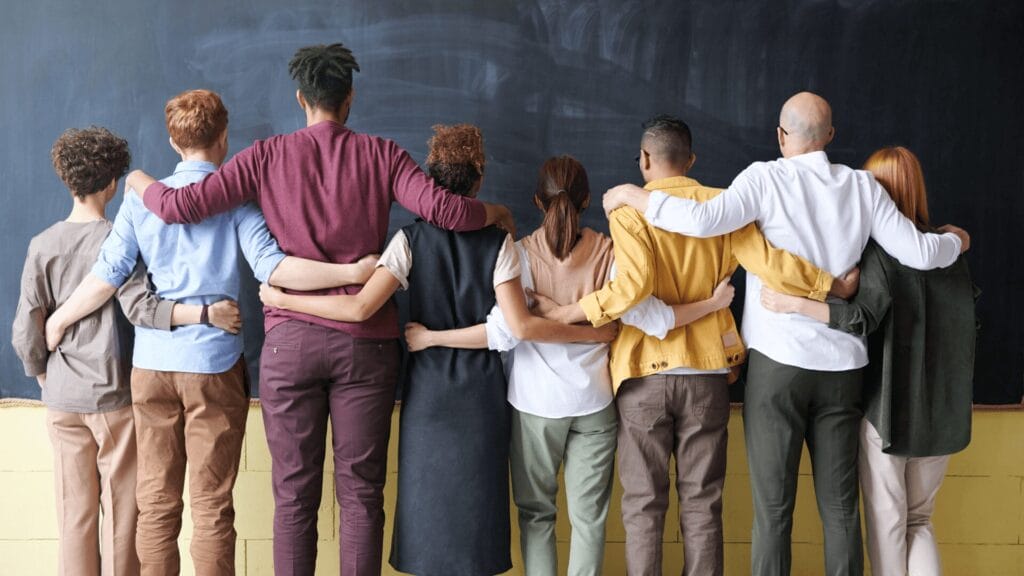 A diverse group of people stands in a line with arms around each other, facing a chalkboard.