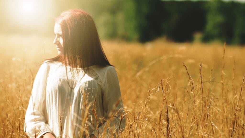 Woman in a white dress stands in a sunny field of tall grass, looking to her left.