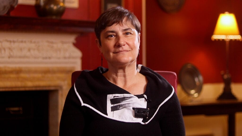 A person with short hair sits in a warmly lit room, wearing a black top with a white and black design.