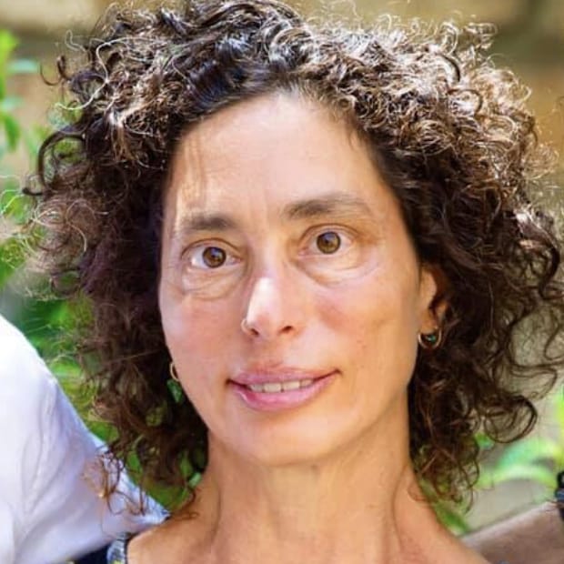 A woman with curly hair and a slight smile poses outdoors with greenery in the background.