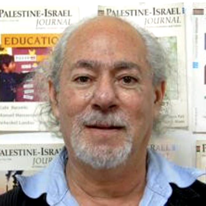An older man with gray hair and beard stands in front of Palestine-Israel Journal posters.