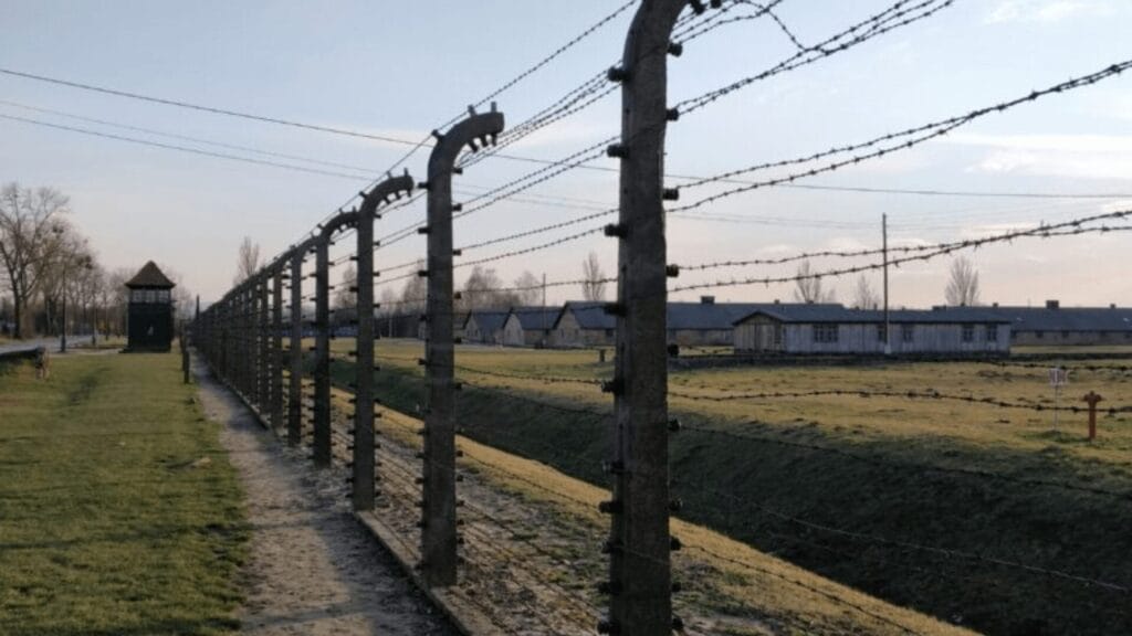 Barbed wire fence and guard tower surrounding a historic concentration camp under a clear sky.