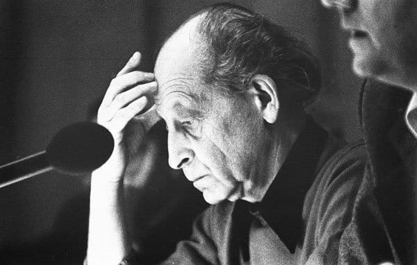 Elderly man sitting at a table, holding his forehead in thought, beside a microphone and another person.