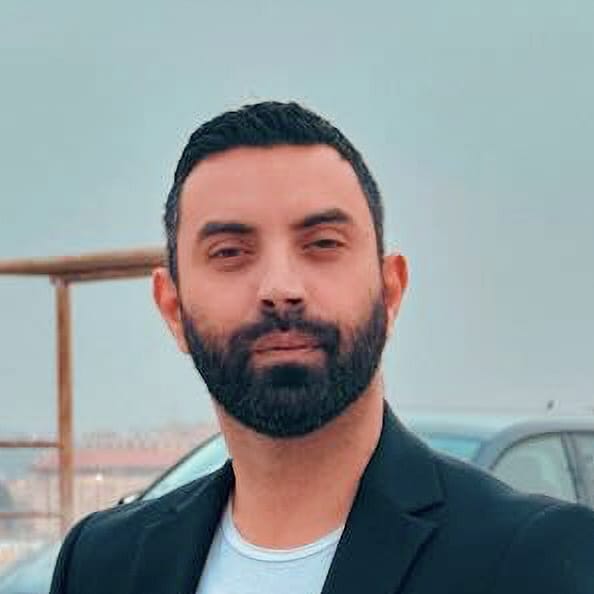 A man with dark hair and a beard wearing a black blazer stands outdoors near a parked car.