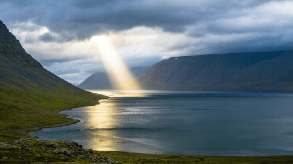 A ray of sunlight breaks through clouds, illuminating a serene lake surrounded by mountains.