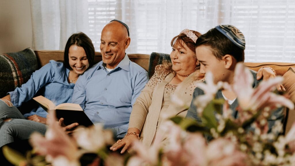 A Jewish family of four sits closely on a couch, smiling and reading a Hagaddah together, with flowers in the foreground.