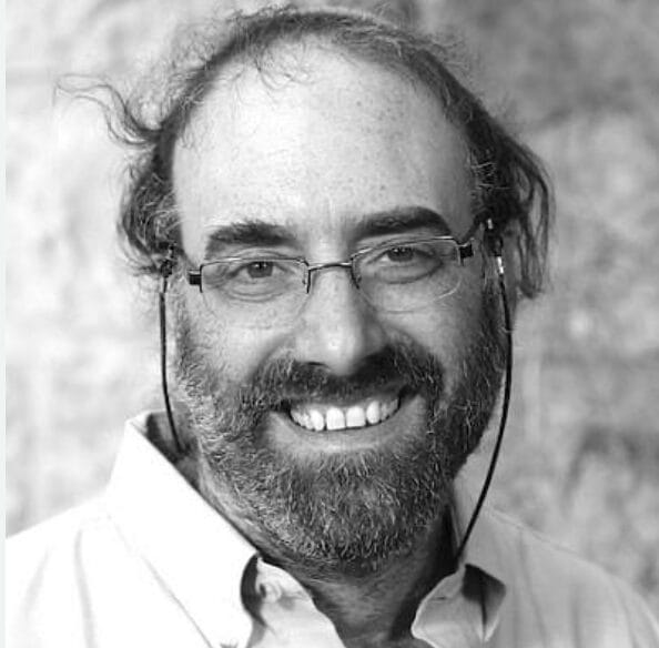 Smiling man with glasses, a beard, and messy hair, wearing a collared shirt, in black and white.