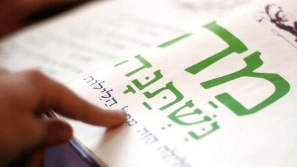 A hand points to Hebrew text in a book with large green letters on a white page.