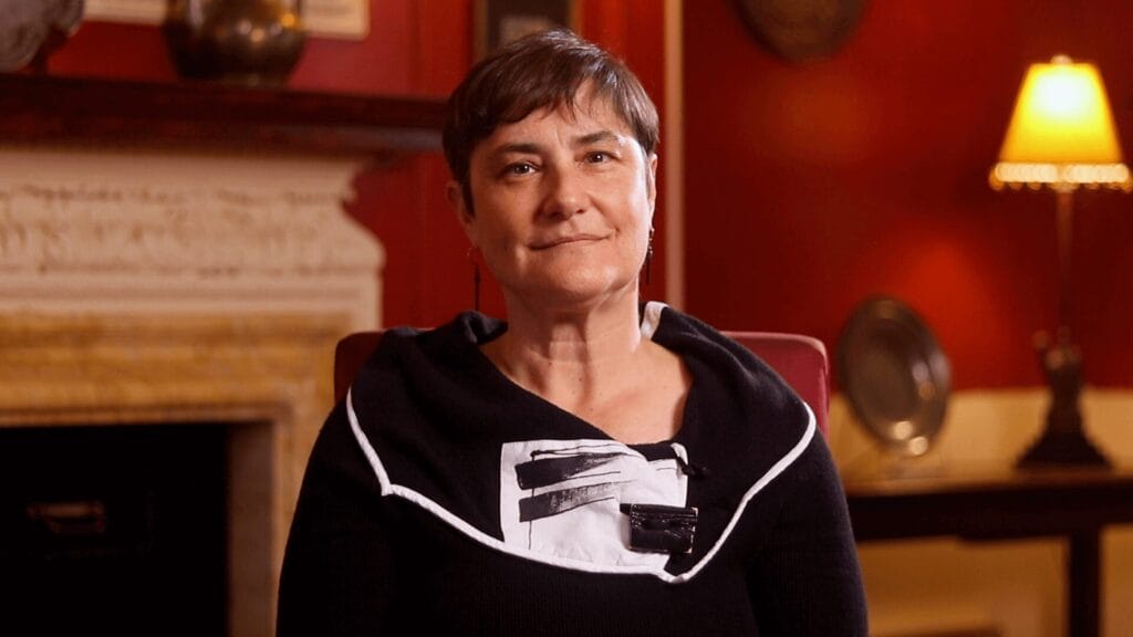 Rabbi Deborah Waxman wearing a black outfit with a patterned scarf, sitting in a warmly lit room with red walls and a fireplace.