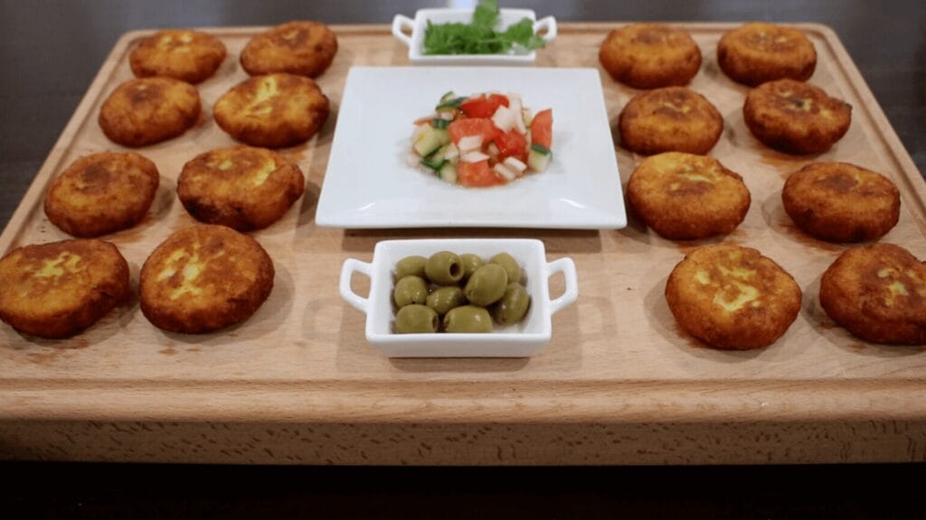 Kubbah potato patties arranged on a wooden board with small dishes of olives and diced vegetables in the center.