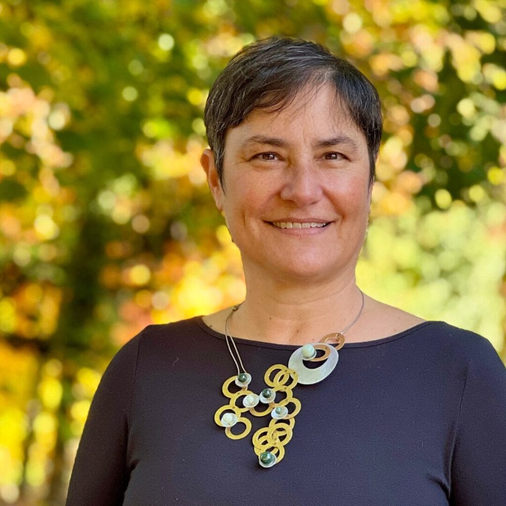Smiling person with short dark hair and statement necklace stands outdoors with a blurred, leafy background.