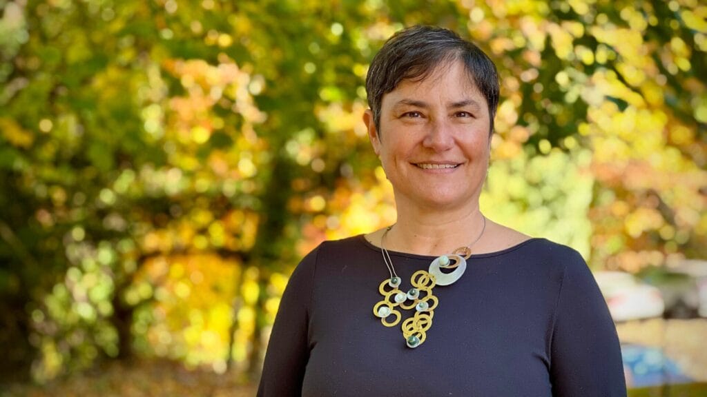 Smiling person with short hair stands outdoors in front of colorful autumn leaves, wearing a dark top and necklace.