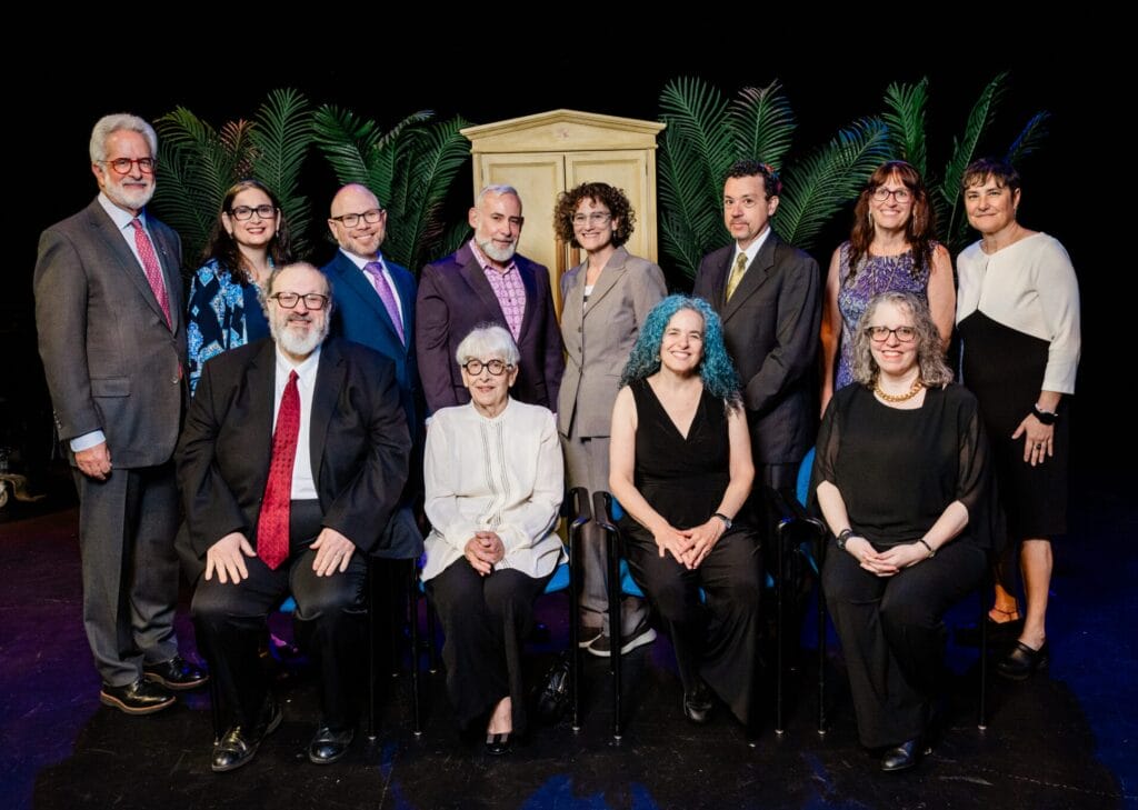 A group of 13 adults pose together on a stage with plants and a cabinet in the background.