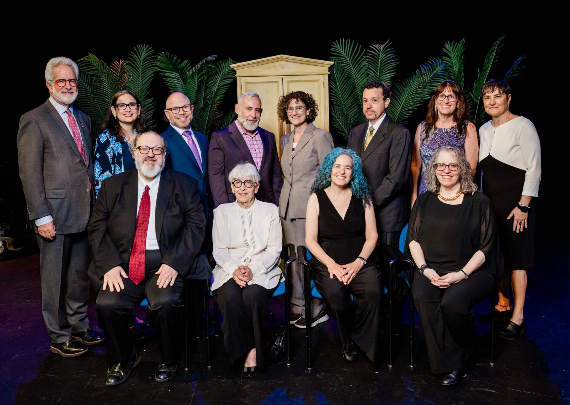 A group of 13 adults pose together on a stage with plants and a cabinet in the background.