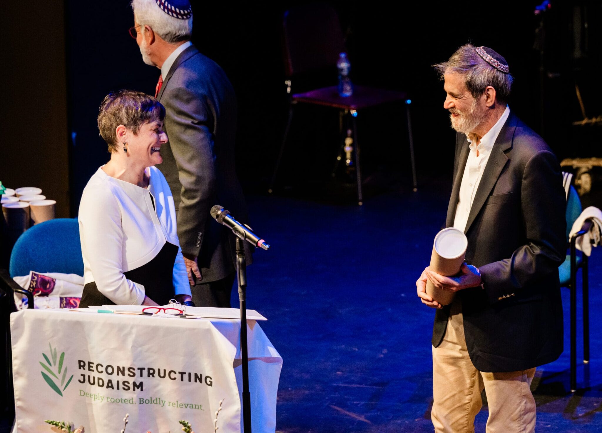 Two people smile and talk on stage at a “Reconstructing Judaism” event, with two others in the background.