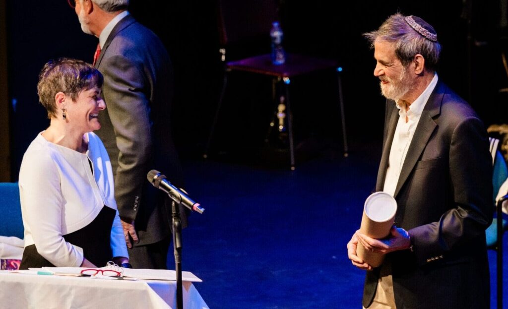 Two people smile and talk on stage at a “Reconstructing Judaism” event, with two others in the background.