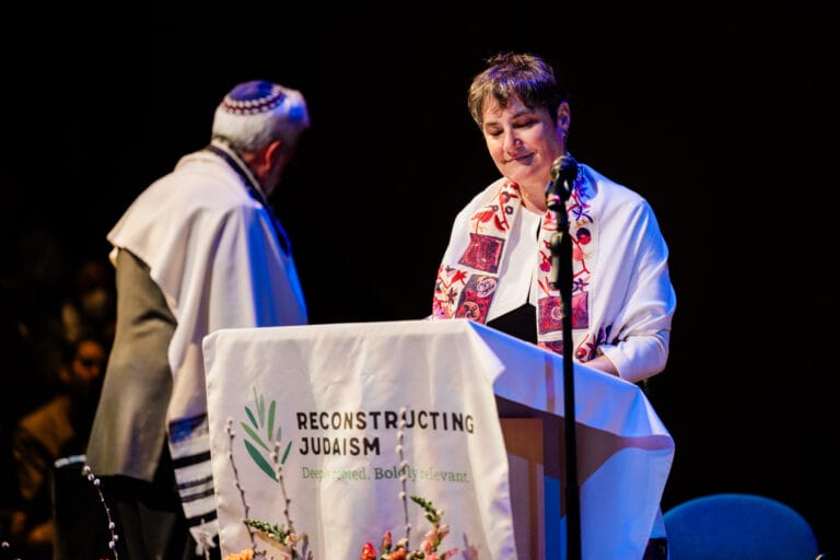 A person in a prayer shawl speaks at a podium labeled Reconstructing Judaism.