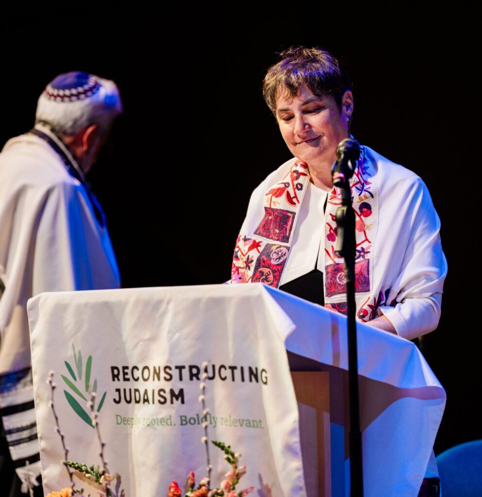 A person stands at a podium reading Reconstructing Judaism while another person stands nearby.
