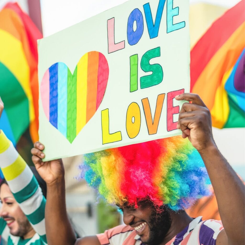 Person in a rainbow wig holds a LOVE IS LOVE sign with a rainbow heart at a Pride event.
