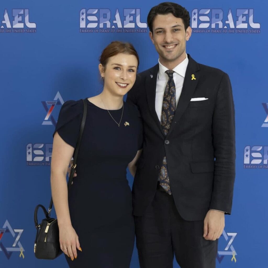 Two people dressed formally stand smiling in front of an Israel embassy backdrop.