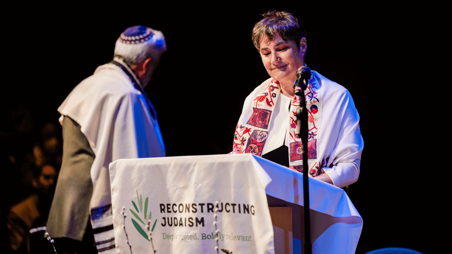 Rabbi Deborah Waxman at a podium reading, with a fellow rabbi, at a Reconstructing Judaism event.