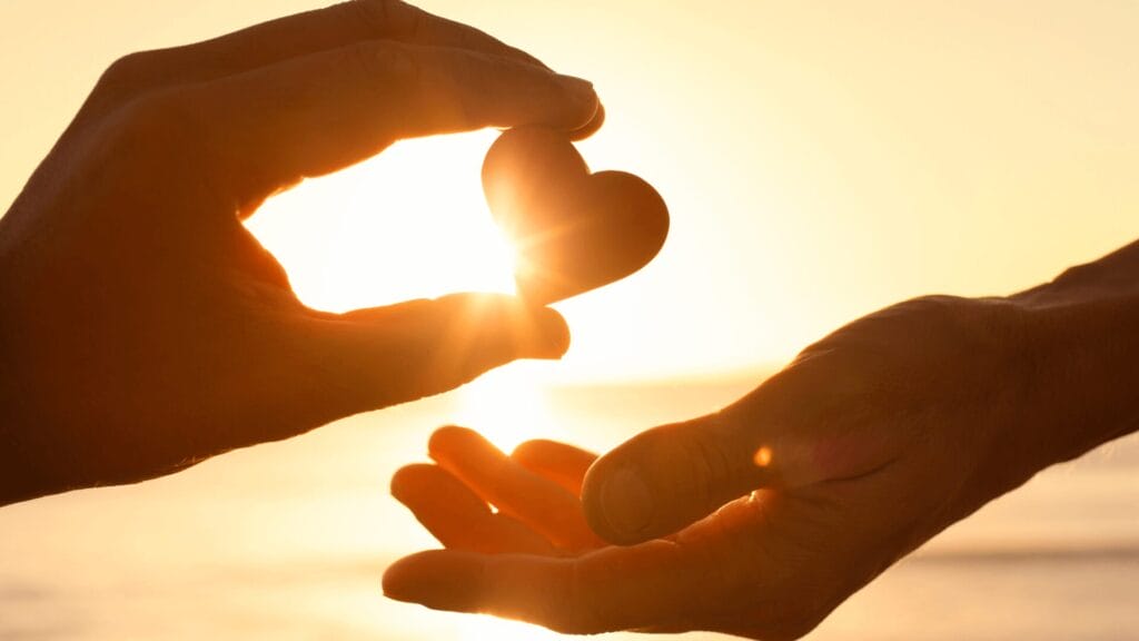 Silhouetted hands exchange a heart-shaped object with sunlight shining in the background.