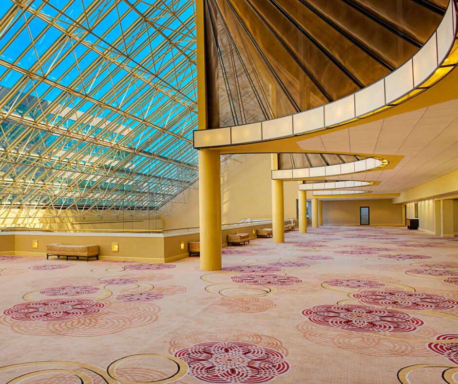 Spacious lobby with patterned carpet, glass walls, and ceiling letting in natural light, with benches along the side.