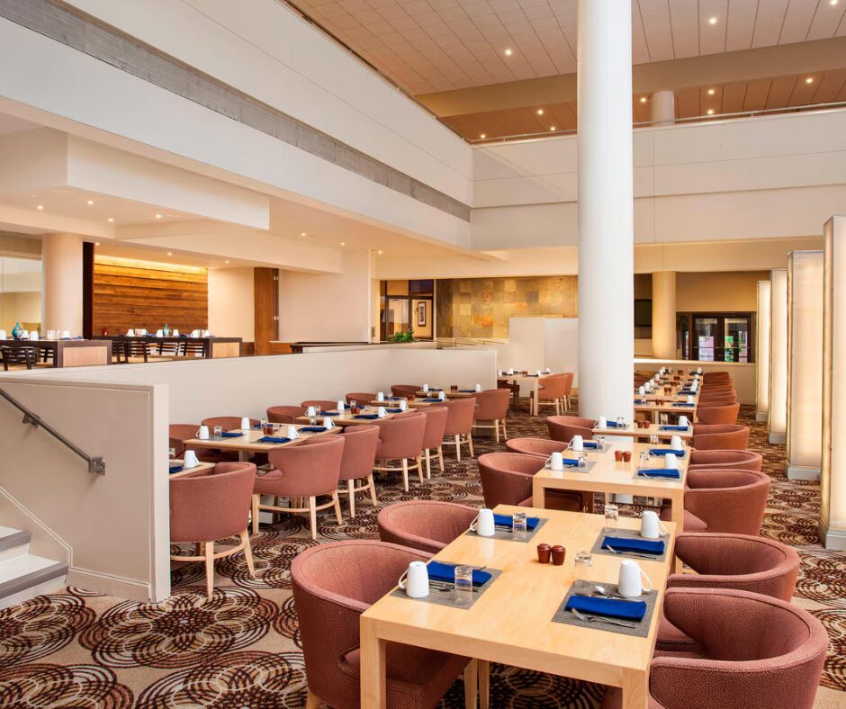 Modern restaurant with wooden tables, pink chairs, and blue napkins, set for dining in a spacious setting.