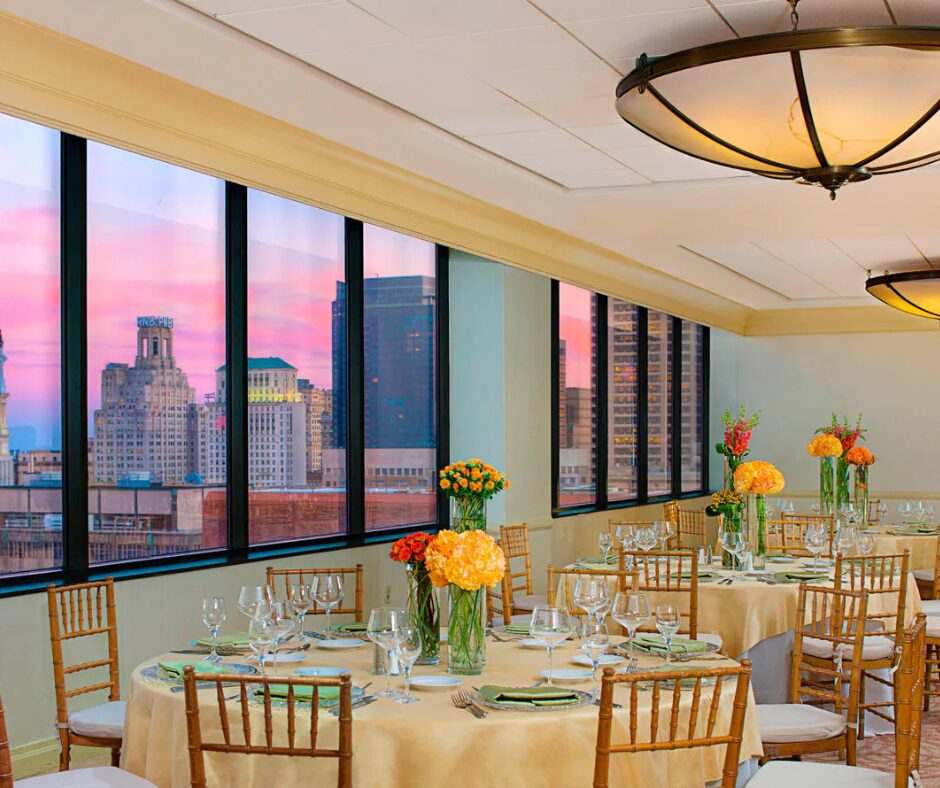 Elegant dining room with round tables, city skyline view through large windows, and colorful flower centerpieces.