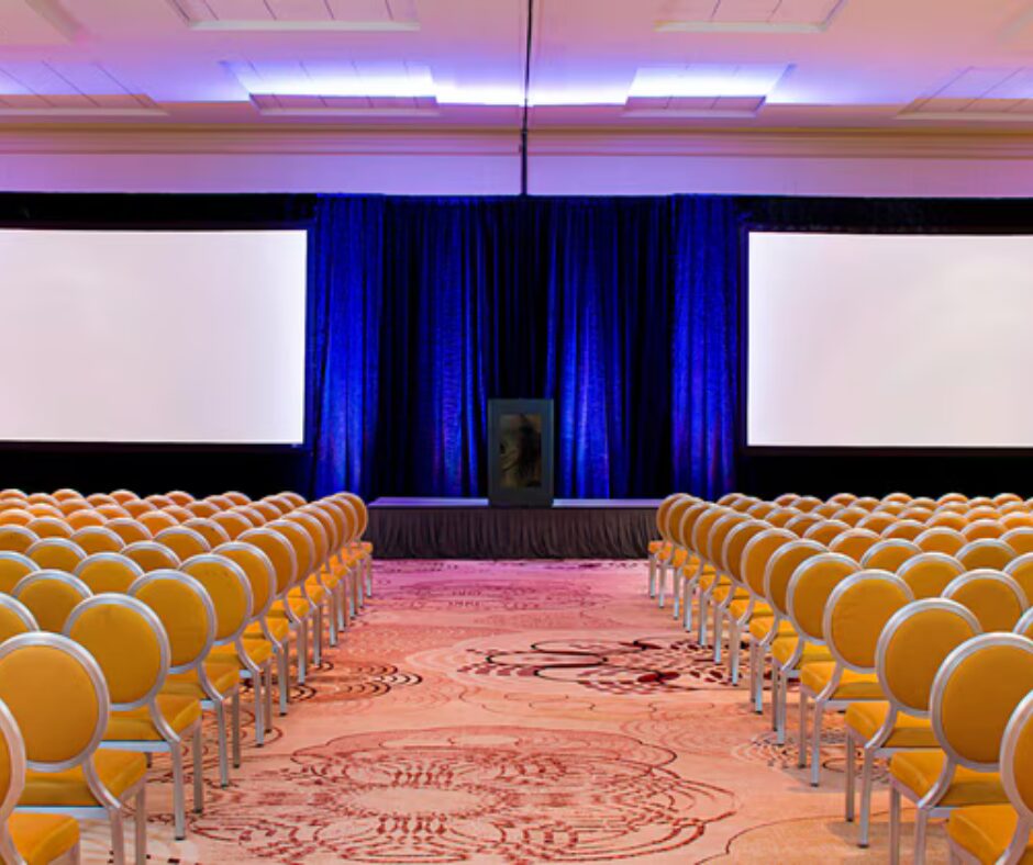 Rows of yellow chairs face a stage with blue curtains and two large projection screens in a conference room.