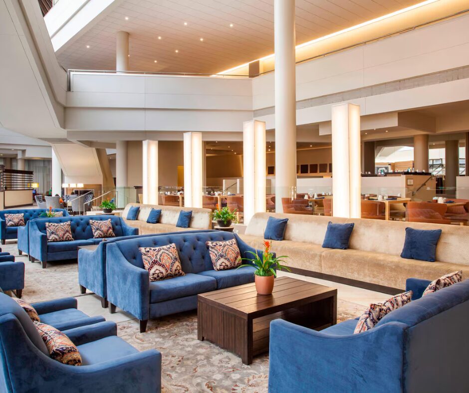Modern hotel lobby with blue and beige sofas, patterned pillows, and potted plants around a wooden coffee table.