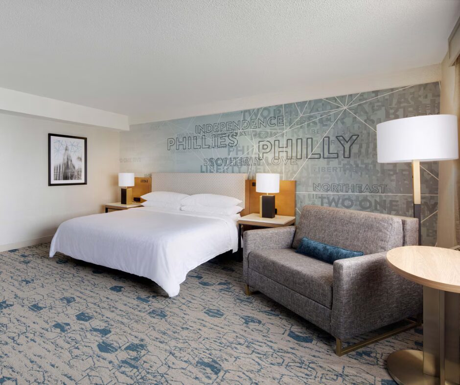 Modern hotel room with a king bed, sofa, lamps, and “Philly” mural on the wall behind the bed.