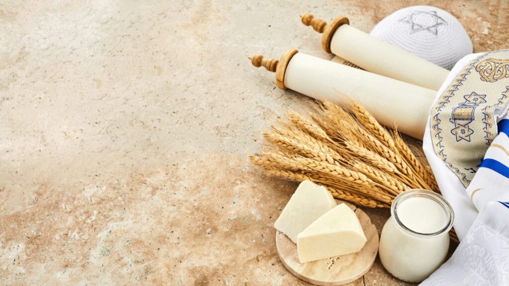 Torah scroll, wheat, cheese, a jar of milk, and a tallit on a light stone surface for Shavuot.