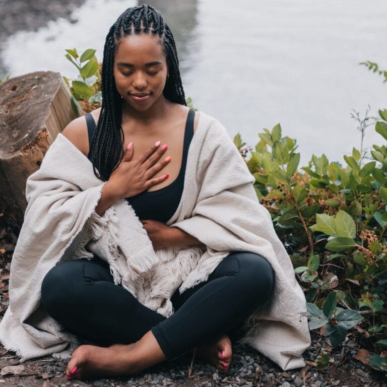 Woman sitting cross-legged by water, eyes closed, hand on chest, wrapped in a blanket, meditating outdoors.