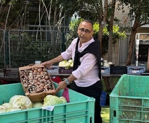 Man holding a box of mushrooms next to large bins filled with cabbages at an outdoor market.