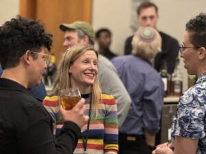 Three people converse at a social gathering, one holding a drink, with others mingling in the background.