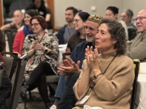 People sitting and smiling at tables, clapping and enjoying an event in a warmly lit room.
